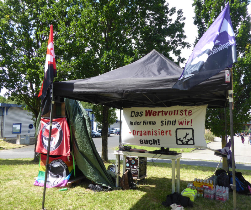 Grüne Wiese. Bäume im Hintergrund. Ein dunkler Pavillion neben einem Turm aus Boxen mit Anti-Flaggen davor. Unter dem Pavillion ein DJ-Pult und ein Banner mit dem Text "Das Wertvollste am Betrieb sind wir - Organisiert euch".